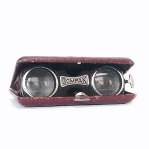 May include: Vintage COMPAK opera glasses in a burgundy leather case. The glasses have a silver-tone metal frame and lenses. The case is open, revealing the lenses. The word "COMPAK" is printed on the front of the case.