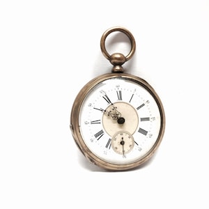 Puede incluir: Un reloj de bolsillo de plata con números romanos en la esfera. El reloj tiene un pequeño dial de segundero y una cadena unida a la parte superior.