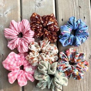 May include: An assortment of fabric scrunchies in various colors and patterns, including pink stripes, gingham, floral prints, and a blue floral design. Some scrunchies have a small tag that reads "be & co."