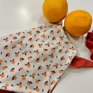 May include: A white bandana with a pattern of orange citrus fruit and green leaves. The bandana has a red trim and the text "bo & co" is printed on the bottom. Two whole oranges are in the background.