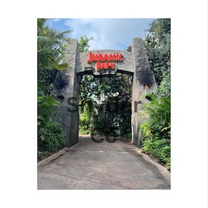May include: A stone archway with the words "JURASSIC PARK" in red letters. The archway is flanked by two large stone pillars and leads to a path surrounded by lush greenery. The sky is visible above the archway.