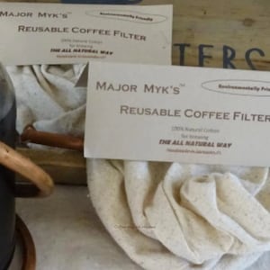 Puede incluir: Un filtro de café reutilizable blanco hecho de algodón 100% natural. El filtro está doblado y colocado sobre una superficie de madera. El embalaje del filtro es visible en el fondo. El embalaje dice "Major Myk's Reusable Coffee Filter" y "The All Natural Way".