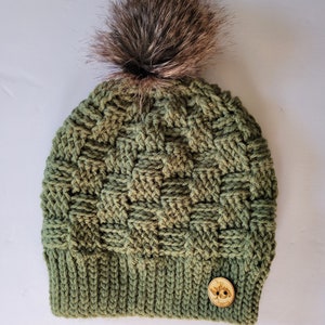 May include: A green crocheted beanie with a brown faux fur pom pom and a wooden button with the text "Tree Hugger" on it.