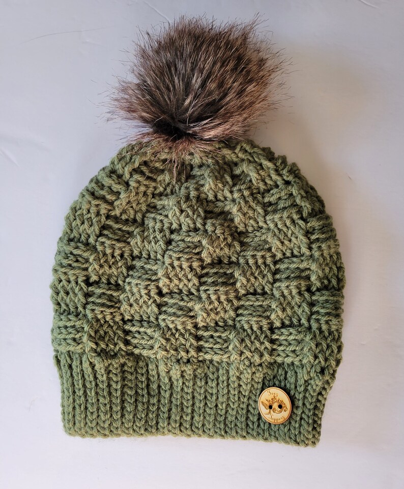 May include: A green crocheted beanie with a brown faux fur pom pom and a wooden button with the text "Tree Hugger" on it.
