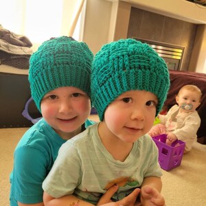 May include: Two children wearing teal knitted hats are sitting together. The child in the front is holding a remote control. The child in the back is wearing a teal t-shirt.
