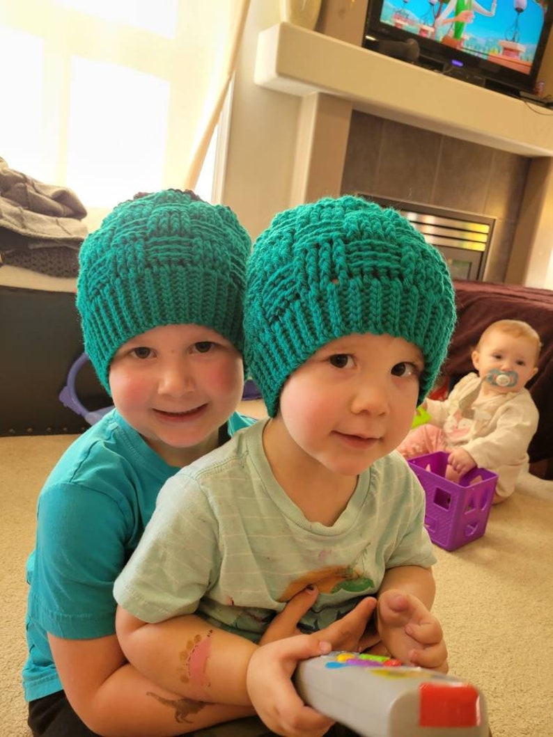 May include: Two children wearing teal knitted hats are sitting together. The child in the front is holding a remote control. The child in the back is wearing a teal t-shirt.