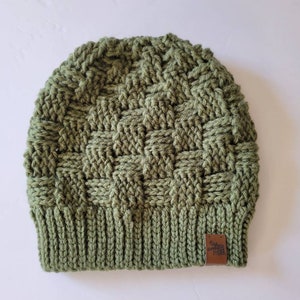 May include: A green crocheted beanie with a brown leather patch featuring a moose design. The beanie has a textured woven pattern.