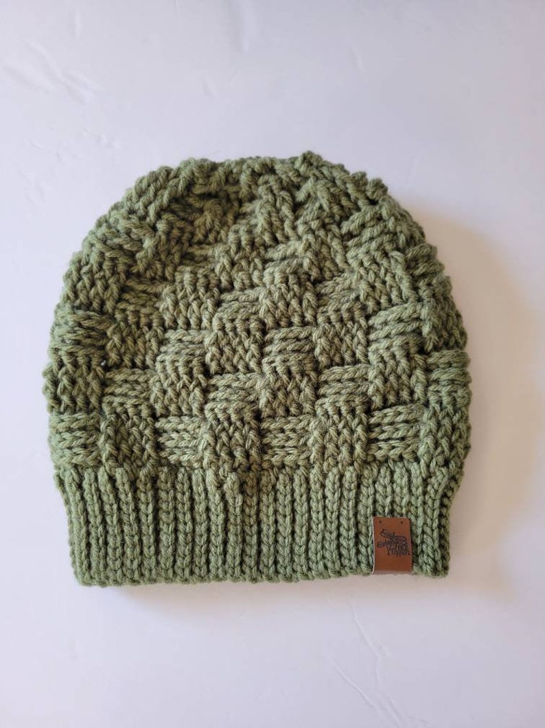 May include: A green crocheted beanie with a brown leather patch featuring a moose design. The beanie has a textured woven pattern.