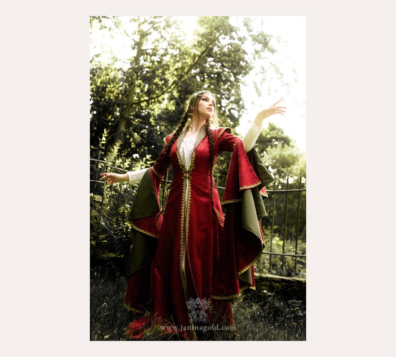 Puede incluir: Un vestido largo rojo de estilo medieval con ribetes dorados y mangas acampanadas. El vestido tiene un vestido interior blanco y detalles en verde oliva en las mangas. La modelo est&aacute; de pie al aire libre con los brazos extendidos. El sitio web www.janinagold.com es visible.