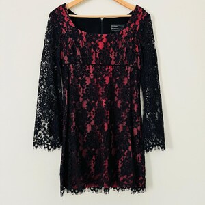 May include: A Nanette Lepore dress featuring a black floral lace overlay on a vibrant pink base. The dress has a square neckline, long lace sleeves with scalloped edges, and a matching scalloped hemline. A black zipper is visible at the center back.