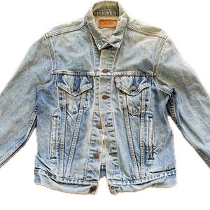 Può includere: Giacca di jeans blu chiaro con bottoni, due tasche sul petto e due tasche laterali. La giacca ha un colletto classico e una piccola etichetta rossa sulla tasca sul petto. Stile vintage.