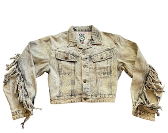 Pode incluir: Uma jaqueta jeans com lavagem ácida clara, apresentando longas franjas desfiadas ao longo das mangas externas. A jaqueta inclui uma gola clássica, fecho frontal com botões, dois bolsos no peito com abas e uma etiqueta no pescoço WOOZ.