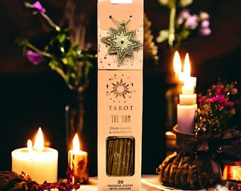 May include: A tall, rectangular light pink box of incense sticks labeled Tarot The Sun Dark Amber & Gingerlily. The box features a gold star-shaped pendant at the top, a sun graphic, and a clear window showing 30 incense sticks with a holder.