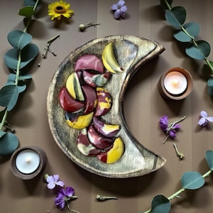 Puede incluir: Una bandeja de madera con forma de media luna contiene varias piedras pequeñas pulidas en forma de media luna en tonos rojo oscuro, amarillo y granate. Dos pequeños portavelas redondos de madera con velas blancas encendidas.