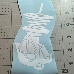 May include: White vinyl sticker of a car suspension coil spring with a shock absorber. The sticker is on a blue background.