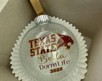 May include: A clear glass ornament with the words "Texas State" in gold and maroon, along with "Bella Dorm Life 2025." The ornament has a gold ribbon and is inside a cardboard box.