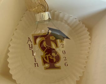 May include: A clear, flat, circular glass ornament featuring a maroon and gold logo with a wildcat silhouette, a black graduation cap, the name Aylin, and the year 2026. The ornament is topped with a silver cap and a gold ribbon for hanging.