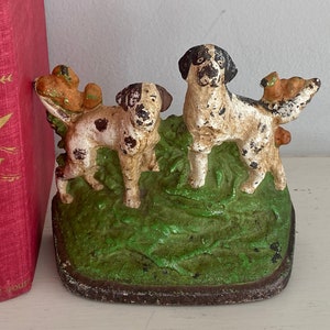 Può includere: Un fermaporta in ghisa con due cani neri e bianchi con macchie marroni, in piedi su una base verde erbosa. I cani tengono piccoli uccelli marroni in bocca.