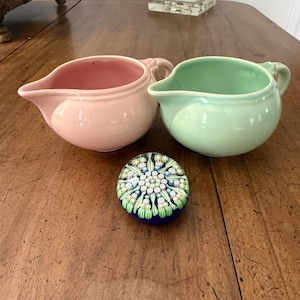May include: Two ceramic cream pitchers, one pink and one mint green, sit on a wooden surface. A round paperweight with a floral design in shades of green, white, and blue is also present. The pitchers have handles and spouts.