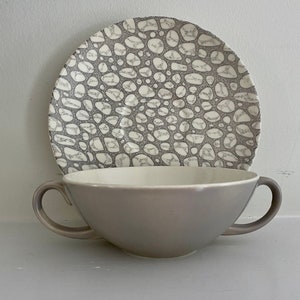 May include: A gray and white ceramic soup bowl with two handles and a matching saucer. The saucer has a pattern of gray and white pebbles.