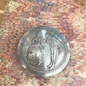 Könnte beinhalten: Ein runder, silberfarbener Metall-Kompaktpuder mit einem detaillierten Relief von zwei Figuren in historischer Kleidung. Der Kompaktpuder liegt auf einem gemusterten Papier mit roten, braunen und cremefarbenen Farbtönen.
