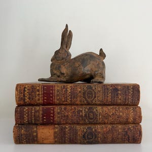 May include: A bronze-colored rabbit figurine rests atop a stack of three antique books. The books have brown leather covers with gold lettering and decorative patterns. The spines of the books read "Percy's Poems".