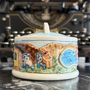 May include: A ceramic lidded jar with a whimsical summer scene. The jar features a blue pool, a brown bunny wearing sunglasses, and two colorful chairs. The jar is decorated with a light blue glaze and has a white base.