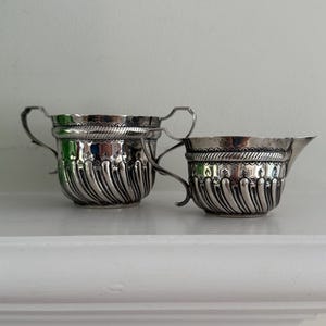 May include: A pair of antique silver serving pieces: a handled sugar bowl and a creamer with a spout. Both items feature a fluted design and decorative trim. The silver reflects light, giving them a bright and elegant appearance. The pieces are likely for tea or coffee.
