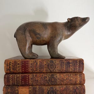 Op de afbeelding: Een bronzen beerfiguur staat op een stapel van drie antieke boeken. De beer heeft een gedetailleerde vachttextuur. De boeken hebben verouderde, getextureerde covers met gouden letters en decoratieve patronen.