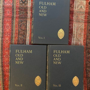 Op de afbeelding: Drie donkerblauwe hardcover boeken met de titel "FULHAM OLD AND NEW" met gouden letters. Elk deel is genummerd met Romeinse cijfers. Een gouden embleem staat op de onderste voorkant van elk boek. De boeken zijn gerangschikt op een patroonstof.