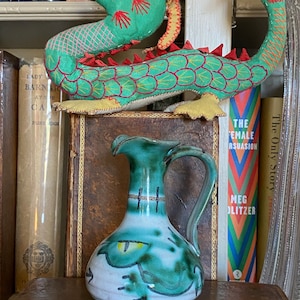 May include: A green and white ceramic pitcher with a handle, decorated with a stylized green eye and a black and white design. The pitcher is sitting on a shelf with a book and a stuffed dragon toy.