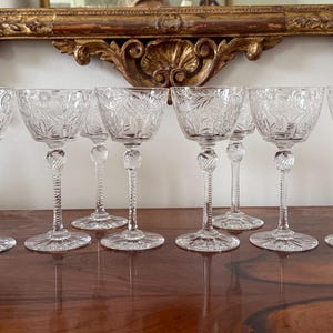 May include: Eight clear glass stemmed goblets with intricate cut designs. The goblets are arranged in a row on a wooden surface.