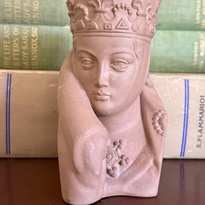 May include: A tan-colored bust of a crowned figure, possibly a queen or saint. The sculpture has a detailed face and is adorned with a crown and ornate details on the garment. The bust is set against a backdrop of vintage books.