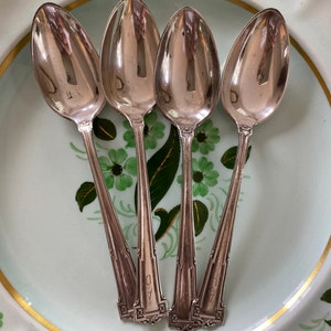 Könnte beinhalten: Vier silberne Löffel mit verzierten Griffen sind fächerförmig auf einem weißen Teller mit grünem und goldenem Blumendesign angeordnet. Die Löffel haben die Nummer "40" auf der Rückseite des Griffs eingraviert.