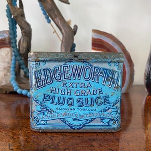 May include: A vintage blue and white tin container for Edgeworth Extra High Grade Plug Slice Smoking Tobacco. The tin has a decorative border and the text "Larus & Bro. Co. Richmond, Va. USA" is printed on the bottom.
