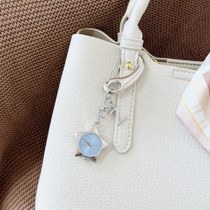 Op de afbeelding: Een crèmekleurige, getextureerde handtas met een zilverkleurige, stervormige horloge-bedel aan het handvat. Een kleine, bedrukte zijden sjaal in beige, bruine en witte tinten is aan de zijkant van het handvat geknoopt als decoratief accent.