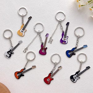 Puede incluir: Una colección de llaveros con forma de guitarra en varios colores, incluyendo amarillo, negro, rojo, morado y azul. Cada llavero tiene un anillo y una cadena de metal. Algunos llaveros tienen colgantes con letras. Los llaveros se muestran sobre una superficie blanca.