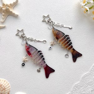 Op de afbeelding: Twee visvormige oorbellen met een stervormige bovenkant, visgratenbedel en kleine schelp- en sterbedels. Eén oorbel is rood, zwart en wit, de andere is bruin en bruin. Op een witte ondergrond met zeesterren en schelpen.