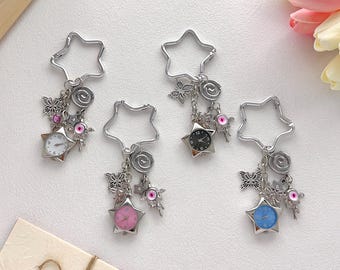 Könnte beinhalten: Vier sternförmige Schlüsselanhänger mit silbernen Metallrahmen. Jeder Schlüsselanhänger hat eine kleine Uhr mit einem unterschiedlich farbigen Zifferblatt: weiß, schwarz, rosa und blau. Sie verfügen außerdem über Schmetterlings- und Blumenanhänger.