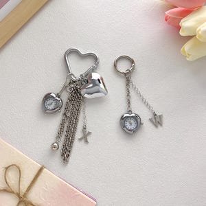 Op de afbeelding: Twee zilveren hartvormige sleutelhangers. Eén sleutelhanger heeft een hartvormig horloge, een hartbedel en een ketting met een X-bedel. De andere heeft een hartvormig horloge en een W-bedel. Op een witte ondergrond.