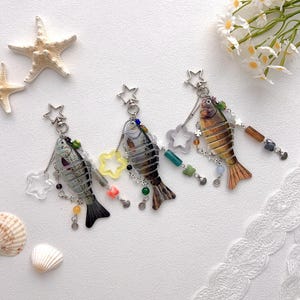 Puede incluir: Tres llaveros con forma de pez con abalorios en forma de estrella y detalles de cuentas. Los peces son en tonos grises, marrones y naranjas, con herrajes de metal plateado. También hay conchas marinas y estrellas de mar decorativas.