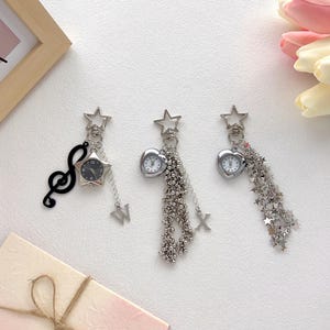 Può includere: Tre portachiavi argentati con piccoli quadranti. Uno presenta un orologio a forma di stella con un ciondolo a forma di chiave di violino. Gli altri due hanno orologi a forma di cuore con catene pendenti e ciondoli a stella. Gli orologi hanno quadranti neri.