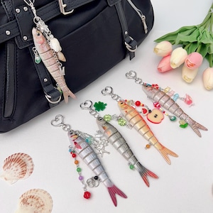 May include: A selection of fish-shaped keyrings in silver, orange, and pink, decorated with beads, charms, and seashells. One keyring is attached to a black handbag. The keyrings are displayed on a white surface.
