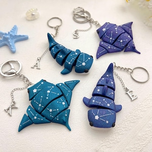 Może przedstawiać: Kolekcja ręcznie robionych breloków do kluczy w niebiesko-białym motywie konstelacji. Breloki przedstawiają różne wzory stworzeń morskich, w tym rekina wielorybiego i manty. Każdy brelok ma srebrny breloczek i zawieszkę z literą.