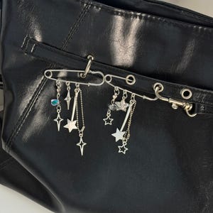 May include: Two silver-toned safety pins attached to a black faux leather bag. Each pin features dangling charms including various star shapes, thin metal rods, small chains, and a single iridescent blue teardrop-shaped bead on the left pin.