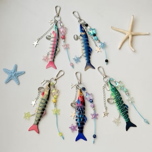May include: Five articulated fish keychains with metal lobster clasps, each featuring colorful patterns, dangling star charms, small seashell accents, and beaded strands. The fish are painted in pink, blue, yellow, orange, and green gradients.