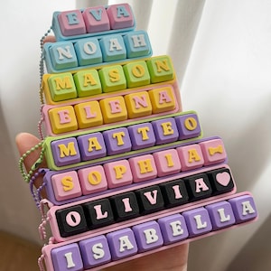 May include: A stack of rectangular keychains featuring colorful plastic keyboard-style keys. The keys display the names EVA, NOAH, MASON, ELENA, MATTEO, SOPHIA, OLIVIA, and ISABELLA in raised letters, with a heart icon on the OLIVIA keychain.
