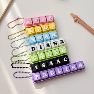 May include: A collection of seven rectangular keychains, each featuring a row of four to seven square keys with letters spelling the names EVIE, CICI, NOAH, DIANA, CALEB, ISAAC, and PAULINA. Each keychain has a matching colored ball chain attachment.