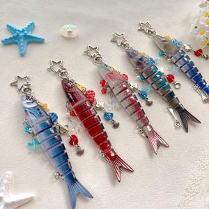 May include: A collection of five fish-shaped keychains in various colors, including blue, red, and gray. Each keychain features a star-shaped clasp and is adorned with decorative beads and charms. The fish have segmented bodies and detailed fins.