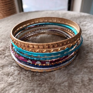 Könnte beinhalten: Ein Stapel bunter Armreifen mit goldenen Akzenten. Die Armreifen sind aus Metall und haben verschiedene Muster und Designs.